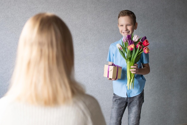 Un garçon mignon dans une chemise bleue donne un bouquet de tulipes et une boîte avec un cadeau à maman