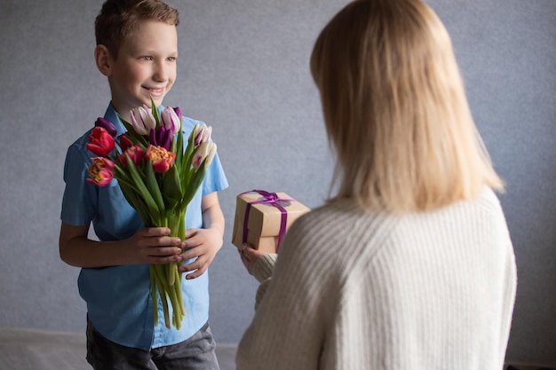 Un garçon mignon dans une chemise bleue donne un bouquet de tulipes et une boîte avec un cadeau à maman