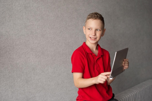 Un garçon mignon dans un t-shirt rouge tient une tablette dans ses mains et regarde ailleurs