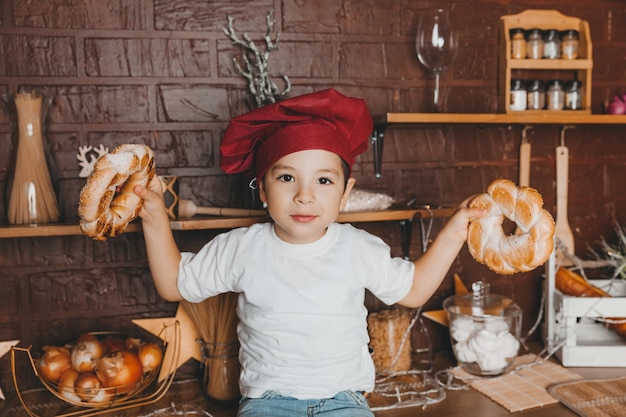 Un garçon mignon dans une toque est assis dans la cuisine et tient des petits pains à bagel près de son visage.