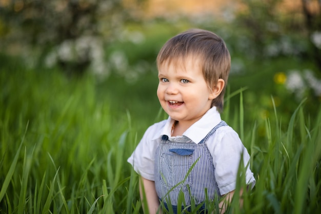 Garçon mignon dans des vêtements à la mode aux yeux bleus joue et saute dans les hautes herbes dans un grand jardin fleuri vert