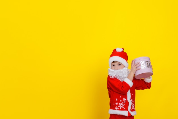 Garçon mignon d'enfant caucasien dans des vêtements de père Noël et avec une barbe a soulevé une boîte-cadeau ronde rose