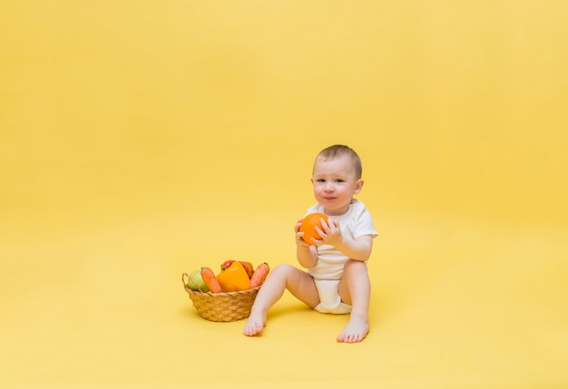 Un garçon mignon est assis dans un body blanc et tient une orange. Un petit garçon est assis avec un panier de légumes et de fruits sur un espace jaune. L'enfant regarde.