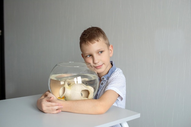 Un garçon mignon est assis près d'un aquarium transparent dans la pièce et embrasse un aquarium avec des poissons