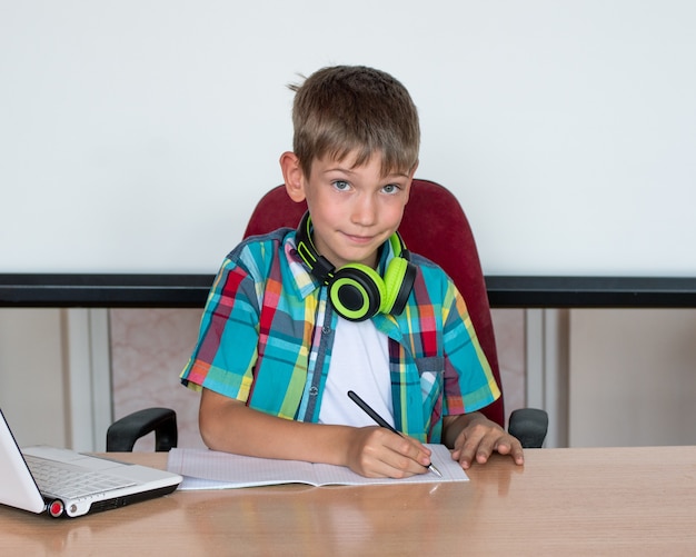 Un garçon mignon est assis à une table, regarde un ordinateur portable, écrit ses devoirs.