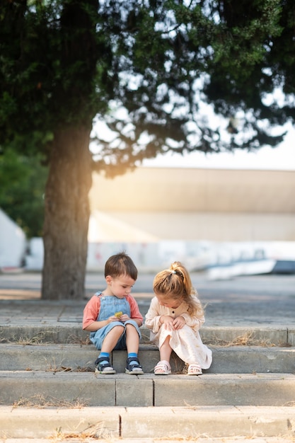 Un garçon mignon et une fille bouclée sont assis sur une marche sous un arbre dans le parc et regardent quelque chose