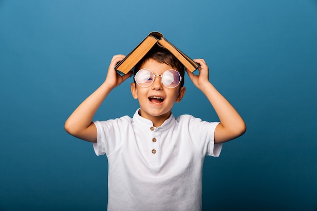 Un garçon mignon avec des lunettes tient un livre sur sa tête, sur fond bleu. garçon avec un livre montre la langue. écolier.