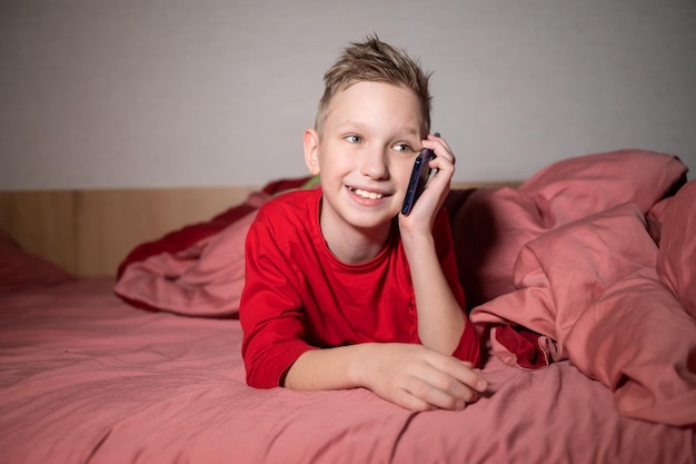 Un garçon mignon en pyjama rouge est allongé dans son lit sous une couverture et parle au téléphone