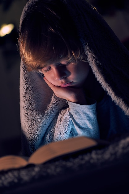 Un garçon mignon qui lit un livre intéressant et s'appuie sur sa main alors qu'il est allongé sur un lit confortable sous une couverture chaude dans une pièce sombre à la maison