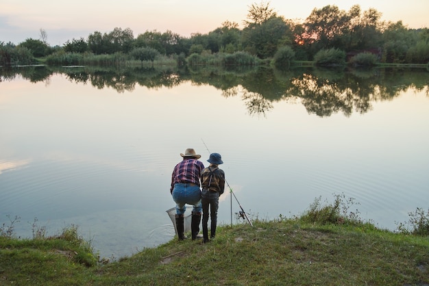 Photo garçon mignon avec son grand-père à la barbe grise attraper des poissons sur le lac avec épuisette au coucher du soleil