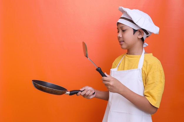 Un garçon mignon souriant portant l'uniforme de chef tient une casserole en téflon et une spatule pour cuisiner