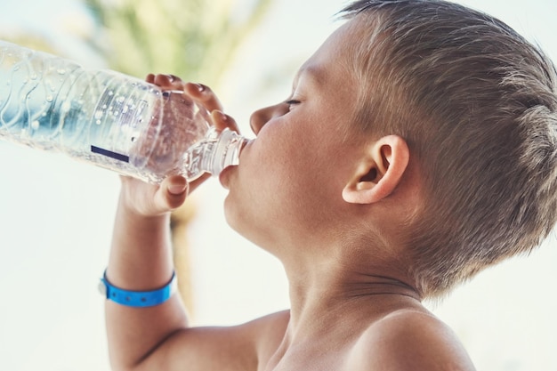 Garçon mince avec torse nu buvant de l'eau minérale en bouteille près de la piscine contre un palmier haut