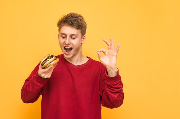 Garçon montre un signe OK et regarde le hamburger dans sa main, isolé sur un jaune