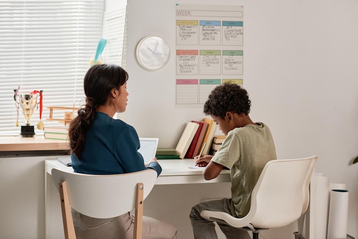 Photo garçon noir aux cheveux bouclés assis à table dans sa chambre avec calendrier sur le mur et faisant des tâches scolaires