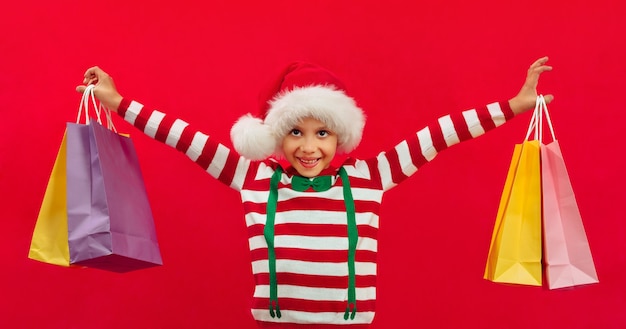 Un garçon noir dans un bonnet de Noel et avec des sacs de transport tient des cadeaux dans ses mains des achats de Noël