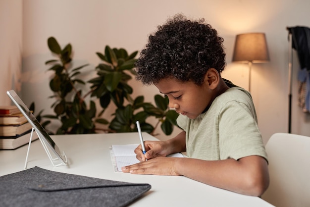 Garçon noir sérieux aux cheveux bouclés assis au bureau et prenant des notes dans le cahier pendant la leçon en ligne à la maison