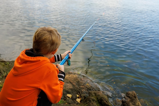 Le garçon pêche avec une canne à pêche sur la rivière tenant une canne à pêche dans ses mains et assis sur les pierres