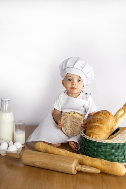 Un garçon un petit cuisinier dans un tablier et une casquette est assis sur une table avec un petit pain dans les mains, du lait, des œufs, du pain