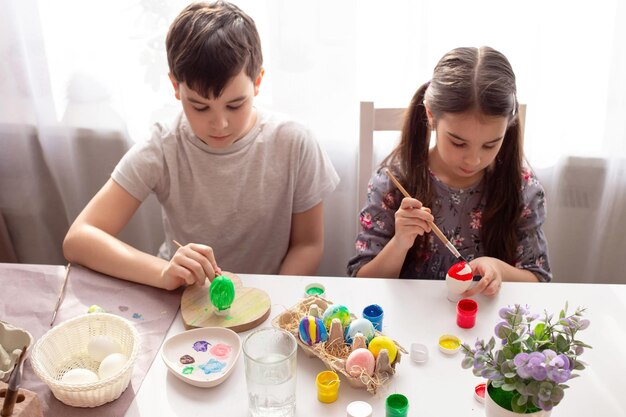 Un garçon et une petite fille sont assis à une table blanche près de la fenêtre se préparent pour les vacances de pâques