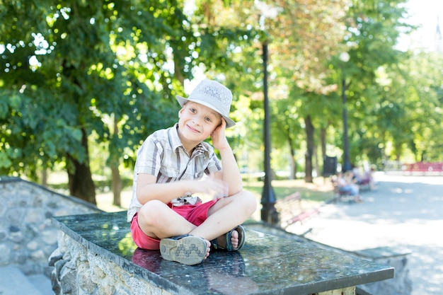 Un garçon portant un chapeau et un short est assis sur un banc de pierre dans un parc.