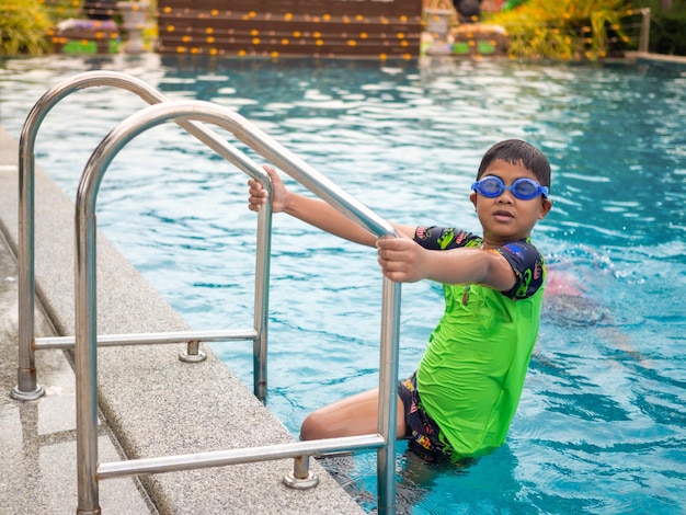 Le garçon portant des lunettes de natation sur l'échelle de la piscine bleu maillot de bain vert