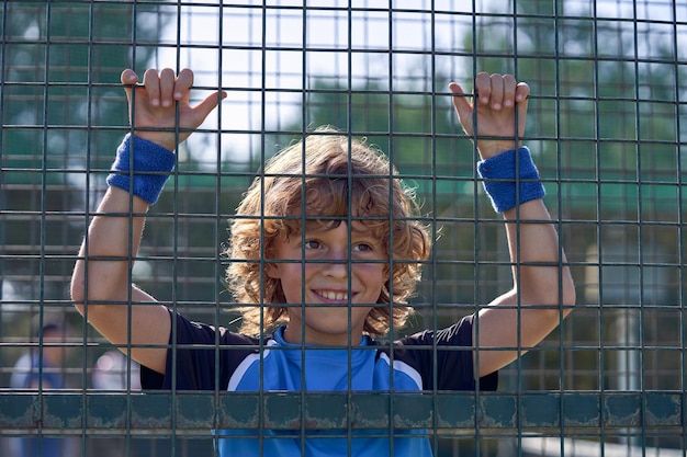 Garçon positif aux cheveux bouclés tenant la main sur un terrain de tennis en métal et regardant ailleurs sur un terrain de sport au soleil