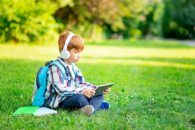 Un garçon de première année avec un sac à dos et une tablette avec des écouteurs sur une pelouse verte lisant un livre ou faisant ses devoirs pour retourner à l'école