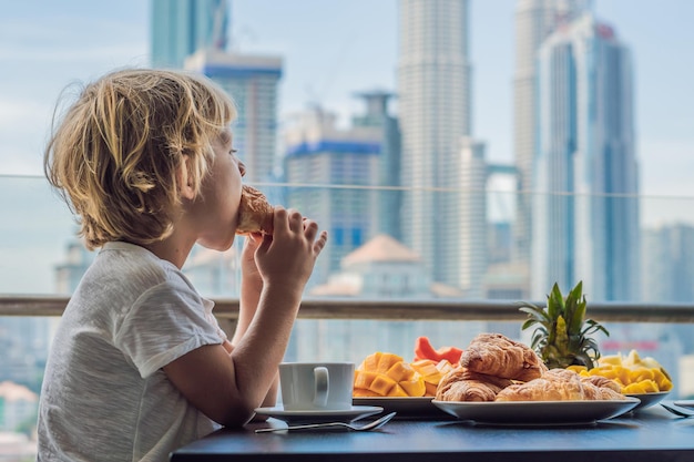 Le garçon prend son petit déjeuner sur le balcon. Table de petit-déjeuner avec café, fruits et pain croissant sur un balcon dans le contexte de la grande ville