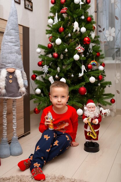 Un garçon en pyjama de Noël tient un Père Noël en chocolat près de l'arbre de Noël à la maison