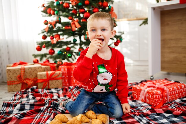 Le garçon qui s'assied près de l'arbre de Noël pour la nouvelle année. Décor de Noël avec des cadeaux, un enfant près du sapin de Noël mange des croissants et sourit