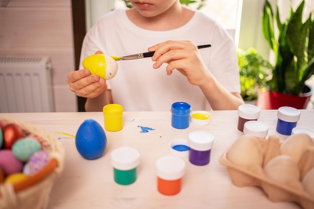 Un garçon de race blanche dans un t-shirt blanc peint des œufs pour célébrer les vacances de Pâques avec des couleurs vives