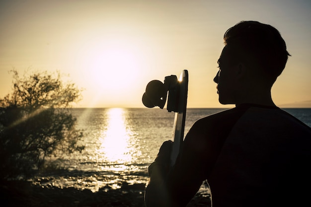 Garçon avec sa planche à roulettes au coucher du soleil assis sur la plage regardant la mer et le ciel - concept de style de vie de liberté - belle adolescente