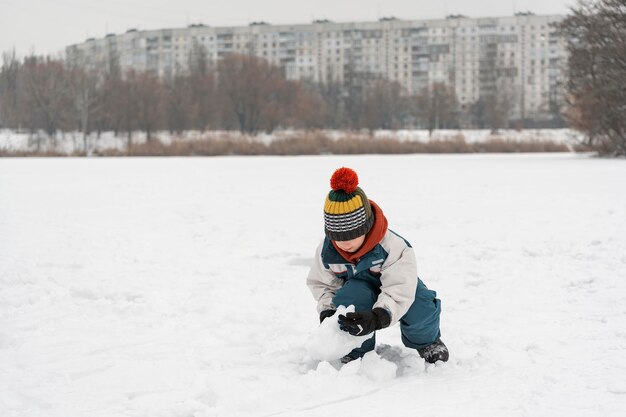 Un garçon sculpte un bonhomme de neige Un enfant joue avec la neige dans un parc d'hiver sur fond d'immeubles de grande hauteur Jeux à l'extérieur en hiver