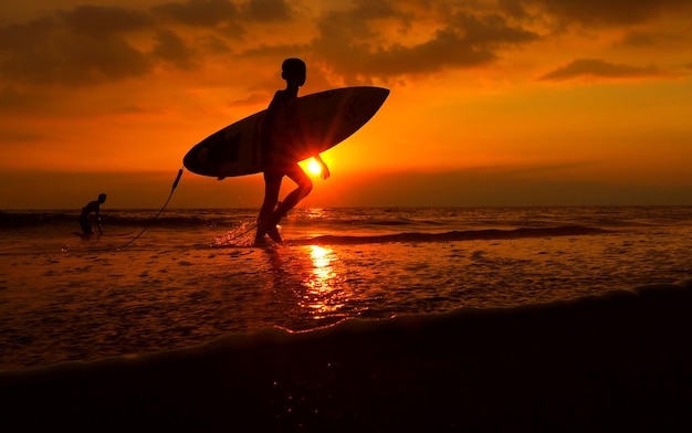 Photo un garçon en silhouette marchant avec une planche de surf en mer au coucher du soleil