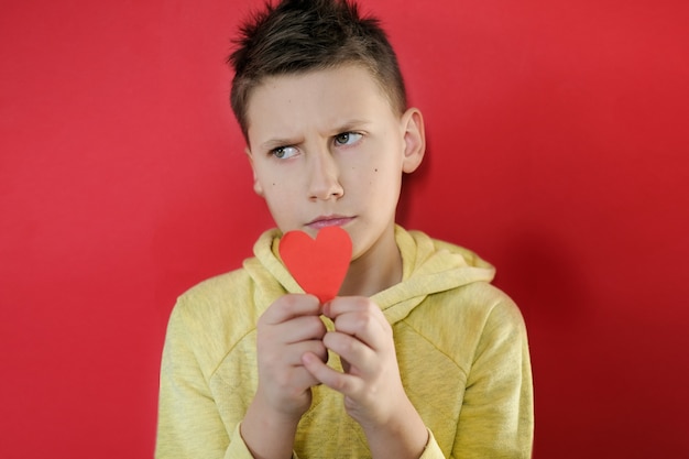 Garçon sombre et en colère en t-shirt jaune montre un coeur de papier rouge au mur rouge