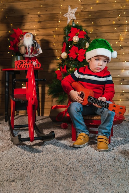 Photo un garçon souriant assis avec une guitare jouet sur le fond du mur en bois et un arbre de noël