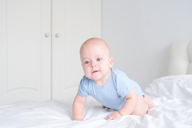 un garçon souriant aux grands yeux bleus dans un body apprend à ramper sur une literie blanche