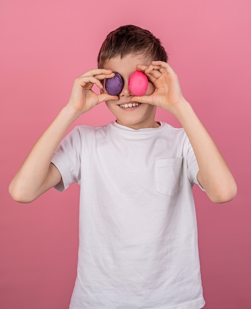 Garçon souriant couvrant les yeux avec des oeufs colorés isolés sur mur rose