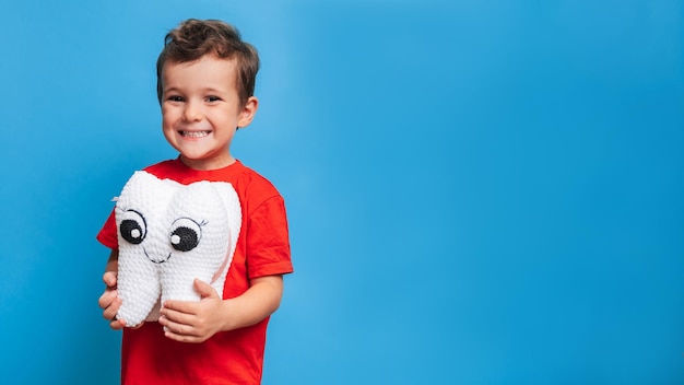 Un garçon souriant avec des dents saines tient une dent en peluche dans ses mains sur un fond bleu isolé Hygiène bucco-dentaire Dentisterie pédiatrique Prévention des caries Une place pour votre texte