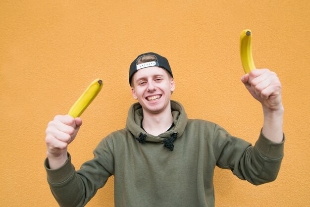 Un garçon souriant est debout sur un mur orange avec des bananes dans ses mains.