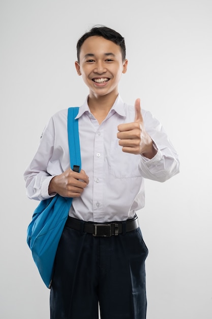 Un garçon souriant portant un uniforme scolaire et portant un sac à dos avec les pouces vers le haut