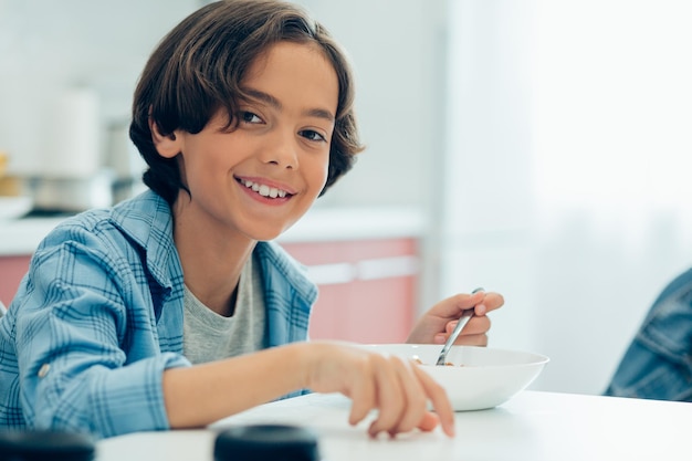 Garçon souriant positif tenant une cuillère assis avec un bol de cornflakes à la table de la cuisine