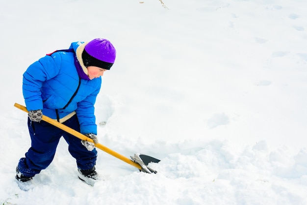 Un garçon souriant transporte de la neige sur une pelle Un enfant nettoie la cour après une chute de neige