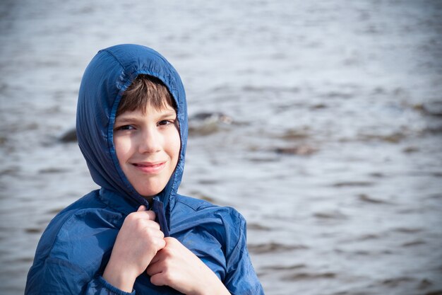 Garçon souriant en veste bleue à capuche sur fond de mer, temps froid.