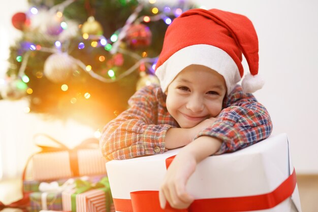 Le garçon de sourire s'asseoir avec une boîte-cadeau près de l'arbre de Noël sur un fond ensoleillé