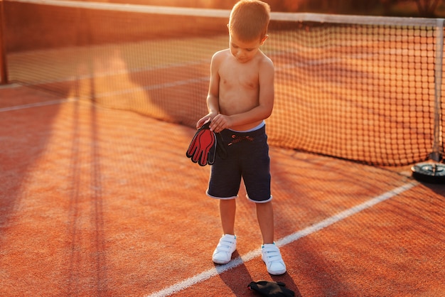 Garçon sportif torse nu mettant des gants de gardien de but en se tenant debout sur le terrain de jeu le matin en été.