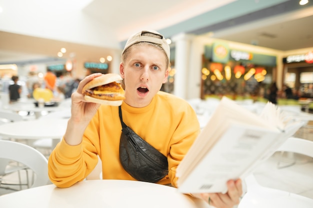 Garçon surpris avec un hamburger et un livre à la main est assis dans un restaurant et regarde la caméra