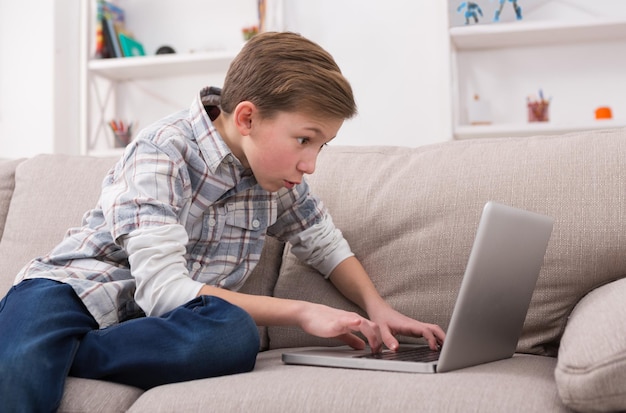 Garçon surpris avec un ordinateur portable sur un canapé à la maison. Enfant en surf occasionnel sur le Web, en tapant sur le clavier