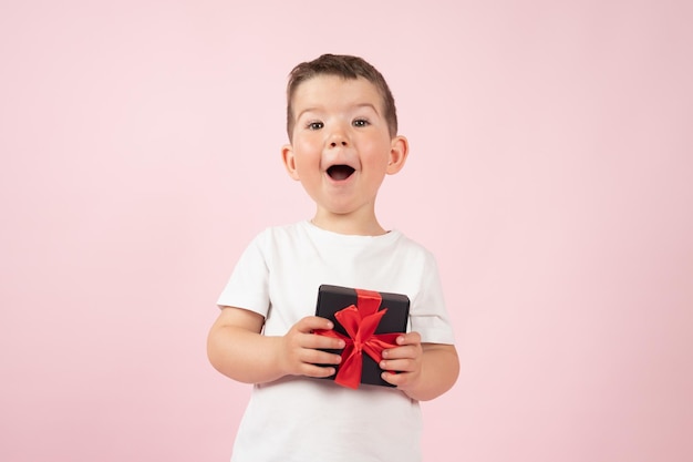 Garçon surpris en t-shirt blanc tenant une boîte cadeau noire devant pour fille ou mère Saint Valentin mère39s jour Fond de studio rose Photo de haute qualité