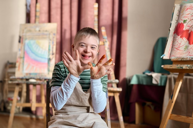 Photo un garçon avec le syndrome de down montrant des mains couvertes de peinture dans un studio d'art